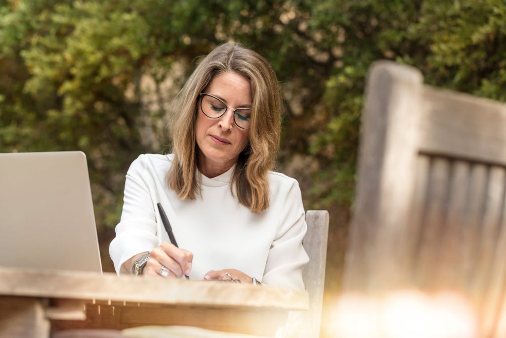 A woman in glasses writing and working on her laptop outdoors in natural daylight.